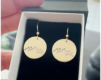 Puede incluir: Un par de pendientes de color dorado con un diseño de cordillera. Los pendientes están en una caja negra.