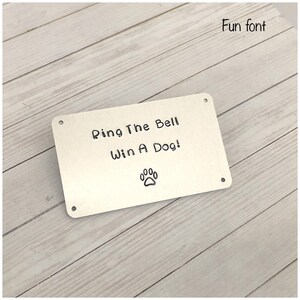 Puede incluir: Una placa rectangular de metal plateado con cuatro peque&ntilde;os orificios en las esquinas para su montaje. La placa est&aacute; grabada con el texto negro "Ring The Bell Win A Dog!" y un peque&ntilde;o icono de huella de pata centrado bajo el texto.