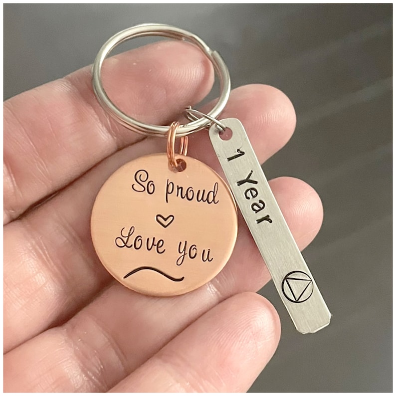 Peut inclure: Un porte-cl&eacute;s en cuivre avec un charme rond qui dit "So proud love you" avec un symbole de c&oelig;ur. Un charme argent&eacute; avec le texte "1 Year" est attach&eacute; au porte-cl&eacute;s.