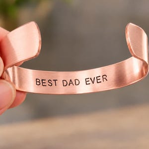 May include: A copper-toned metal cuff bracelet with an open design. The interior surface of the bracelet features the engraved black text BEST DAD EVER centered along the band. The metal has a brushed finish with rounded ends on both sides.