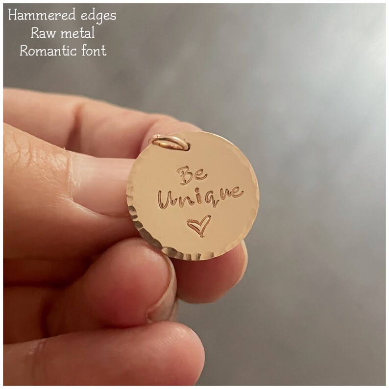 May include: Gold-toned metal charm with a hammered texture. The charm is round and has the words "Be Unique" engraved on it with a small heart symbol.