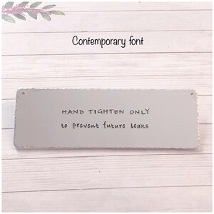 May include: A silver metal sign with rounded edges and holes in the top corners. The sign reads "HAND TIGHTEN ONLY to prevent future leaks" in black handwritten font.