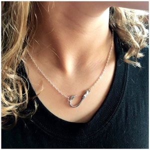 May include: A silver necklace with a fish hook pendant. The necklace is on a model wearing a black shirt.