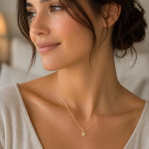 May include: A fine gold necklace with a small, circular pendant bearing a single letter. The pendant is suspended from a slender chain, sitting on the neckline of a person wearing a light-coloured top. The necklace is a simple and elegant piece of jewellery.