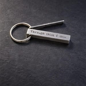 May include: A silver-toned metal keychain featuring a rectangular bar pendant engraved with the text "Through thick & thin" attached to a circular split ring. The keychain has a smooth, matte finish and a simple, functional design.