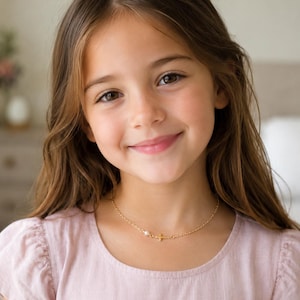 Puede incluir: Un collar de cadena dorada con un pequeño colgante de cruz y una perla blanca como acento. La delicada cadena de metal descansa alrededor del cuello, mostrando el diseño minimalista de la cruz y la perla redonda colocadas una al lado de la otra.