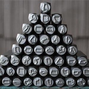 Puede incluir: Una colecci&oacute;n de herramientas de estampado de metal de color plateado dispuestas en forma de pir&aacute;mide. Cada herramienta presenta una forma en relieve &uacute;nica en su extremo plano, dise&ntilde;ada para imprimir patrones. Las herramientas tienen una secci&oacute;n transversal cuadrada.