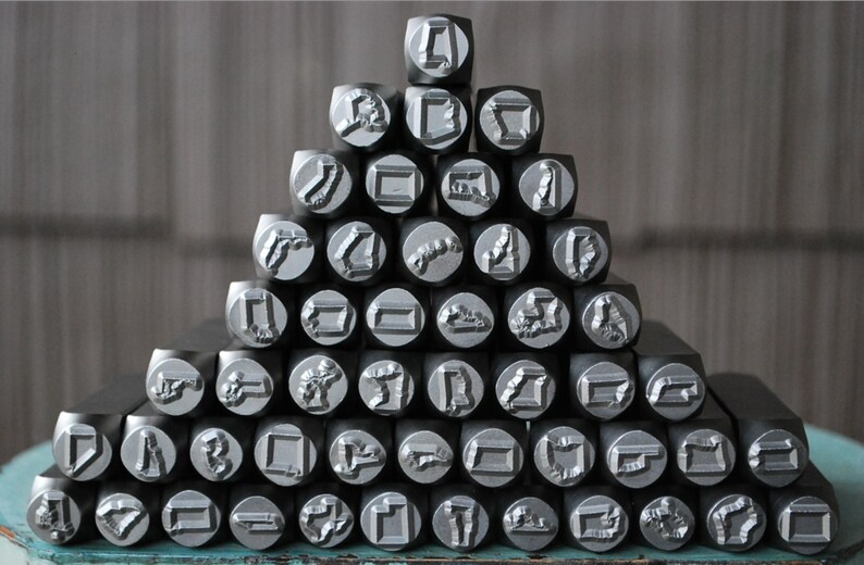 May include: A pyramid-shaped stack of silver-colored metal stamping tools, each featuring a unique embossed design on the flat circular face. The tools are arranged in rows, with a single tool at the top and a wider base of ten tools at the bottom.