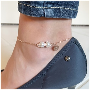 May include: A delicate gold anklet with two white pearls and a small heart charm. The anklet is worn on a person's ankle.