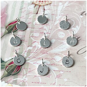 May include: Nine silver charms with different letters on them. The letters are G, L, A, C, B, and J. The charms are on a pink and white background.