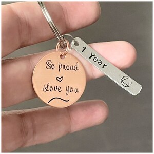 Peut inclure: Un porte-cl&eacute;s en cuivre avec le texte "So proud love you" et un symbole de c&oelig;ur. Une &eacute;tiquette argent&eacute;e attach&eacute;e au porte-cl&eacute;s indique "1 Year".