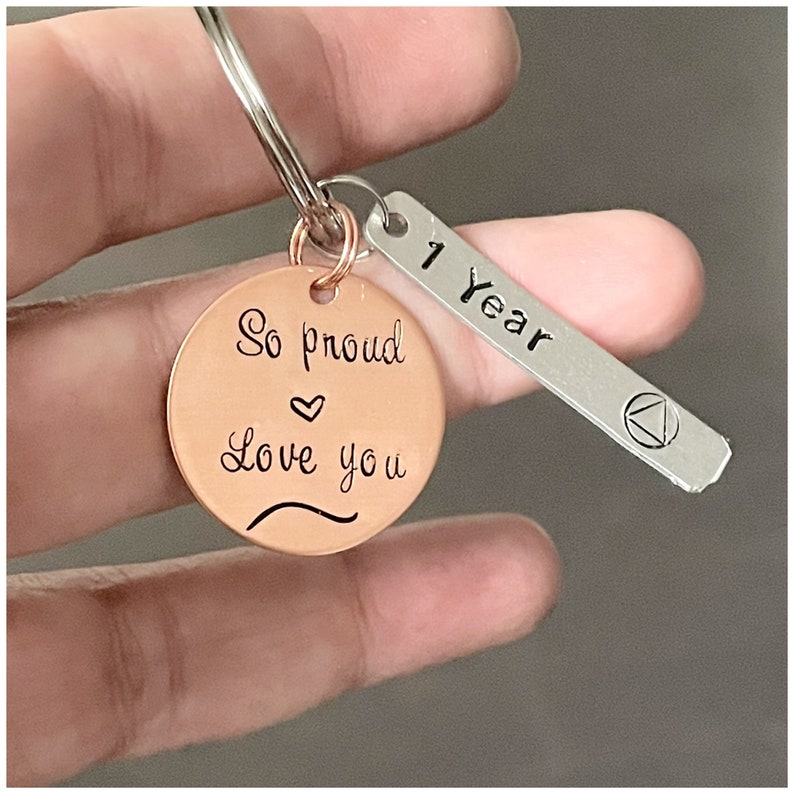 Peut inclure: Un porte-cl&eacute;s en cuivre avec le texte "So proud love you" et un symbole de c&oelig;ur. Une &eacute;tiquette argent&eacute;e attach&eacute;e au porte-cl&eacute;s indique "1 Year".