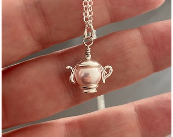 May include: A silver-toned necklace featuring a miniature teapot pendant. The teapot has a pink pearl-like center and detailed handle and spout. The chain is a delicate silver link design, creating an elegant and charming accessory.