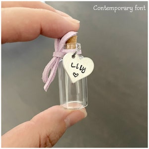 May include: A small glass bottle with a cork stopper and a pink ribbon. A silver heart charm with the name "Lily" is attached to the bottle.