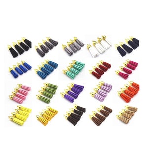 May include: A set of 24 small tassels in various colors, including black, white, gray, blue, green, pink, purple, red, orange, yellow, brown, and beige. Each tassel has a gold metal loop at the top.