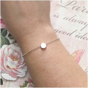 May include: A delicate silver chain bracelet with a small, round silver charm. The bracelet is on a person's wrist.