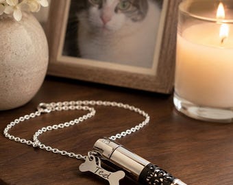 May include: A silver-toned cylindrical pendant necklace featuring a black filigree section and a small bone-shaped charm engraved with the name Ted. The pendant hangs from a delicate silver-toned chain, creating a memorial piece for a pet.