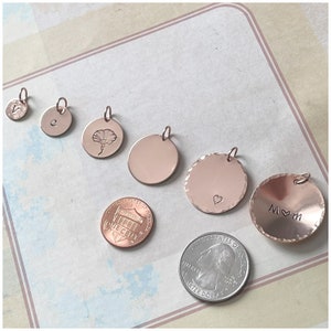 May include: A collection of six rose gold circular charms of varying sizes, some featuring engraved letters, a flower, a heart, or the text M heart M. A US one-cent coin and a US quarter coin are placed nearby for scale reference.