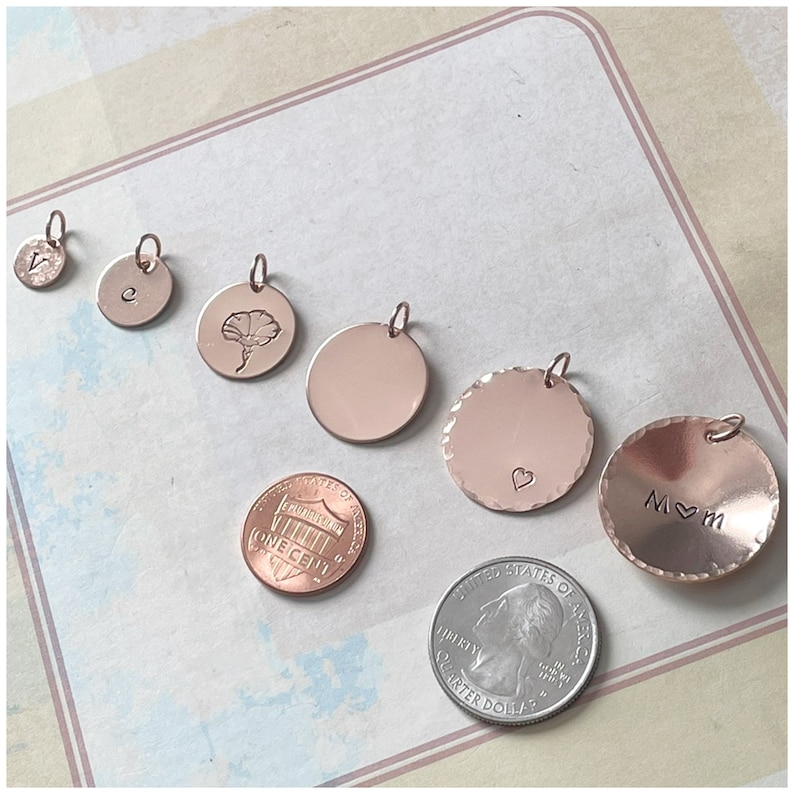 May include: A collection of six rose gold circular charms of varying sizes, some featuring engraved letters, a flower, a heart, or the text M heart M. A US one-cent coin and a US quarter coin are placed nearby for scale reference.