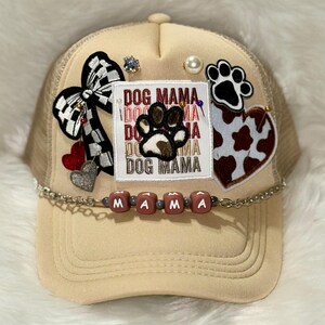 May include: A beige trucker cap with a mesh back, decorated with a black and white chequered bow, a heart, and a patch that reads "DOG MAMA" with a paw print. The brim has the word "MAMA" in beads.