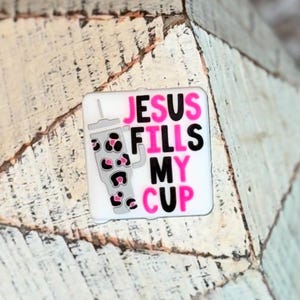 May include: A white square with the text "JESUS FILLS MY CUP" in black and pink. A gray cup with leopard print accents is on the left side of the square. The background is a white and gray wood grain.