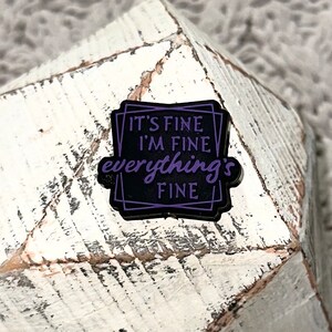 May include: A black and purple enamel pin with the text "It's fine I'm fine everything's fine" in a white font.