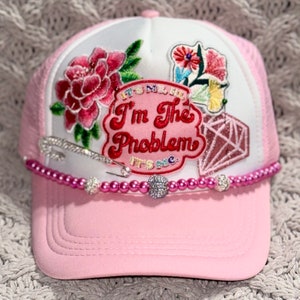 May include: Pink and white trucker hat with a pink diamond, a flower, and a patch that says "I'm the problem it's me" in red and white lettering. The hat has a pink beaded trim with silver safety pins.