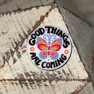 Peut inclure: Un bouton rond blanc avec l'inscription "GOOD THINGS ARE COMING" en noir. Un motif de papillon coloré avec des ailes roses, oranges, jaunes et bleues est au centre. Deux cercles oranges sont de chaque côté.