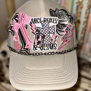 Può includere: Un cappello da camionista grigio con una varietà di toppe, tra cui una croce rosa con il testo "Juice Boxes & Jesus" e una croce a scacchi in bianco e nero.