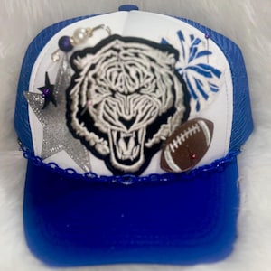 May include: A blue and white trucker hat with a white tiger, a silver star, a football, and blue and white pom-poms. The hat has a blue mesh back and a blue brim.