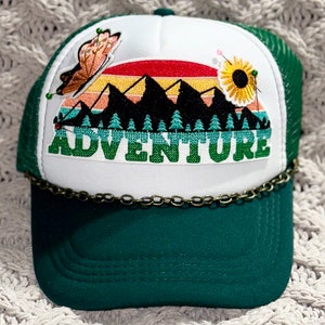 May include: A white and green trucker hat with a chain detail. The front of the hat features an embroidered patch with a mountain range, a sunset, a butterfly, a sunflower, and the word "ADVENTURE".