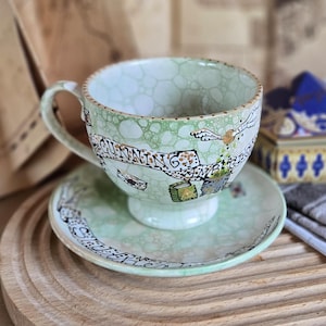 Könnte beinhalten: Ein Keramik-Tee-Set mit einer hellgrünen, blasenartigen Musterung. Die Tasse hat einen Henkel und die Worte "Cunning, Ambitious" auf einem Banner. Die Untertasse hat einen passenden Text am Rand. Das Set steht auf einem Holztablett.