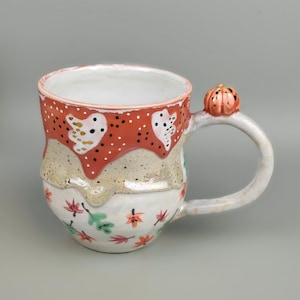 Könnte beinhalten: Eine handgefertigte Keramiktasse mit einem einzigartigen Design. Der obere Teil ist rot mit weißen Punkten und geisterhaften Formen. Der mittlere Teil ist beige, und der Boden weist Blumen- und Blättermuster auf. Ein kleiner Kürbis ziert den Griff.