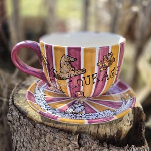 Könnte beinhalten: Ein Keramik-Teebecher- und Untertassen-Set mit einem gestreiften Design in Orange- und Burgundertönen. Die Tasse zeigt das Wort "COURAGE" und Illustrationen eines Hut und einer Kreatur. Die Untertasse hat ein passendes Design.