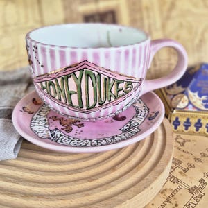 Könnte beinhalten: Ein rosa Tasse-und-Untertasse-Set mit vertikalen Streifen und dem Text "HONEYDUKES" in Diamantenform. Die Untertasse hat ein passendes Design mit einem Frosch und Text. Das Set steht auf einem Holztablett.