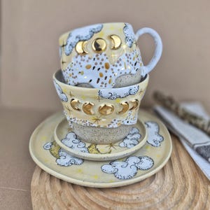 Könnte beinhalten: Ein Stapel handgefertigter Keramikgeschirr, darunter eine Tasse, eine Schüssel und zwei Teller. Die Teile sind mit einem Himmelsmotiv bemalt, das goldene Mondphasen, Wolken und Sterne auf gelbem Hintergrund zeigt. Die Tasse hat einen weißen Griff.