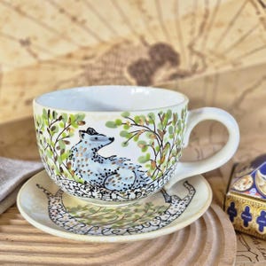 Könnte beinhalten: Cremefarbenes Keramik-Tee-Set. Die Tasse zeigt ein handgemaltes Dachs-Design mit grünem Laub und goldenen Akzenten. Die Untertasse spiegelt das Design der Tasse wider. Das Set steht auf einem Holztablett.