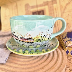 Könnte beinhalten: Ein hellgrünes Keramik-Tee-Set. Die Tasse und die Untertasse sind mit einer Zugszene und den Worten "All aboard the Hogwarts Express" verziert. Die Tasse hat einen Henkel.