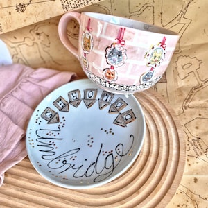Könnte beinhalten: Ein rosa Keramikbecher und -tellerset. Der Becher zeigt Katzenillustrationen und die Worte "I MUST". Der Teller trägt die Worte "The Bridge" und "SHORE". Das Set befindet sich auf einem Holztablett mit Kartenhintergrund.