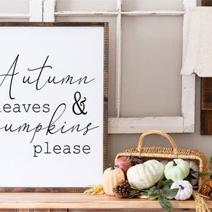May include: A white sign with black calligraphy that reads "Autumn leaves & pumpkins please". The sign is leaning against a white window frame. A wicker basket filled with decorative pumpkins sits on a wooden table in front of the sign.