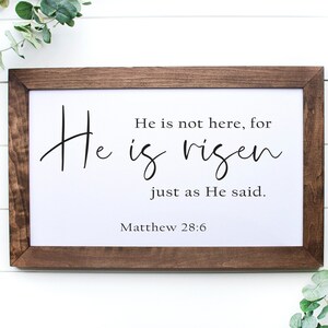 May include: A framed print with a white background and black text that reads "He is not here, for He is risen just as He said. Matthew 28:6". The frame is brown wood.