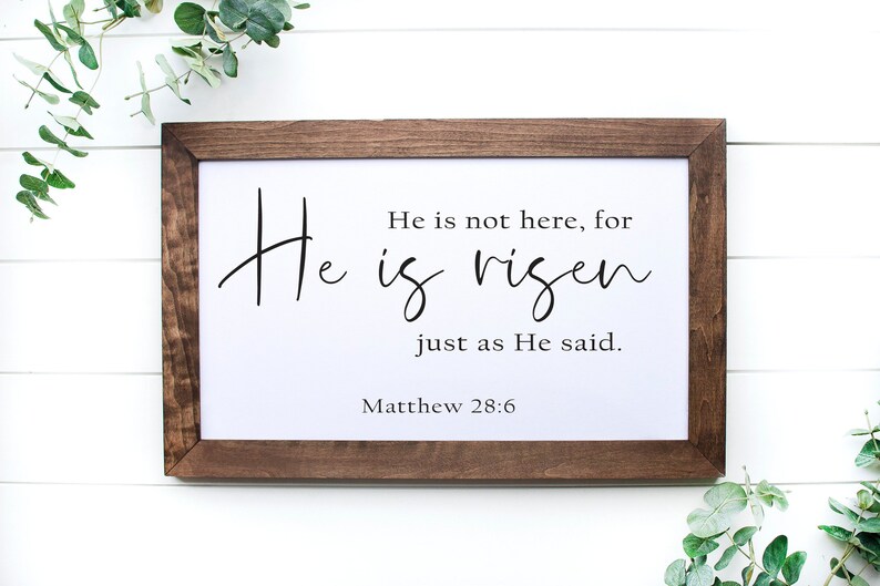 May include: A framed print with a white background and black text that reads "He is not here, for He is risen just as He said. Matthew 28:6". The frame is brown wood.