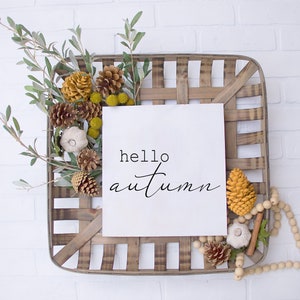 May include: A wooden basket filled with fall decor, including pine cones, gourds, and greenery. A white card with the text "hello autumn" is displayed in the center of the basket.