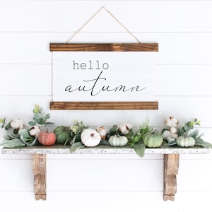 May include: A white sign with the text "hello autumn" hanging on a white wall above a shelf with green leaves, white cotton balls, and small green and orange pumpkins.