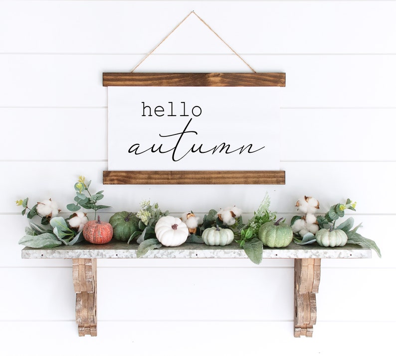 May include: A white sign with the text "hello autumn" hanging on a white wall above a shelf with green leaves, white cotton balls, and small green and orange pumpkins.