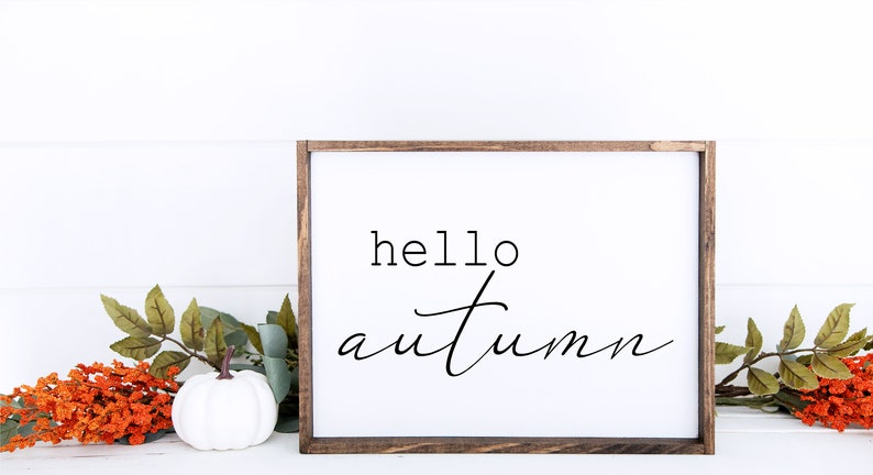 May include: A white wooden sign with a brown frame, featuring the text "hello autumn" in black script font. The sign is sitting on a white surface with a small white pumpkin and orange and green floral accents.
