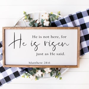 May include: A wooden sign with a white background and black text that reads "He is not here, for He is risen just as He said. Matthew 28:6". The sign is sitting on a white wooden surface with a white and black checkered cloth and cotton flowers.