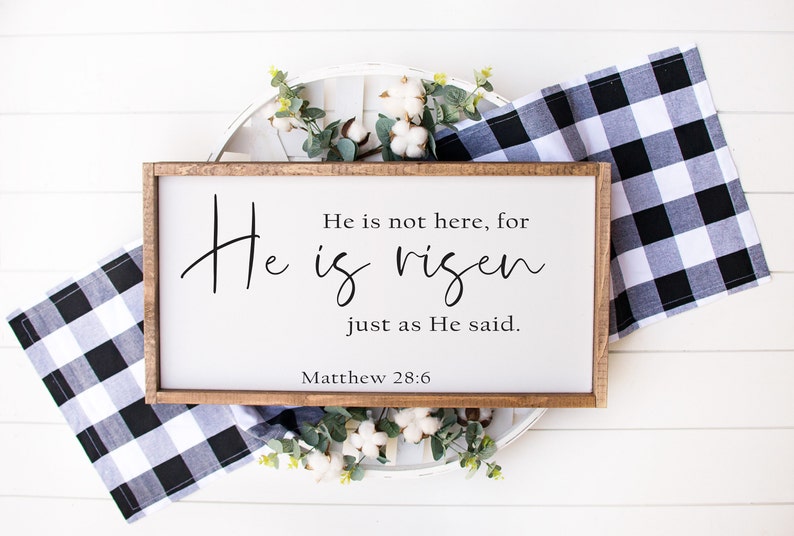 May include: A wooden sign with a white background and black text that reads "He is not here, for He is risen just as He said. Matthew 28:6". The sign is sitting on a white wooden surface with a white and black checkered cloth and cotton flowers.