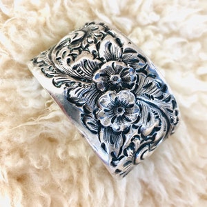 May include: A silver-toned, rectangular belt buckle with an ornate floral design. The buckle features detailed carvings of flowers and leaves, creating a textured surface. The buckle is set against a soft, white, fluffy background.