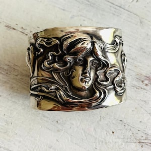 May include: A wide, silver-toned cuff bracelet. The bracelet features a detailed relief of a woman's face with flowing hair and floral accents. The design is Art Nouveau in style, with a polished finish.